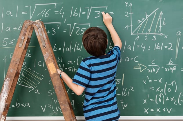 Highly intelligent little boy in the classroom standing on a stepladder to reach a complex mathematical problem on the blackboard that he is busy solving.jpeg